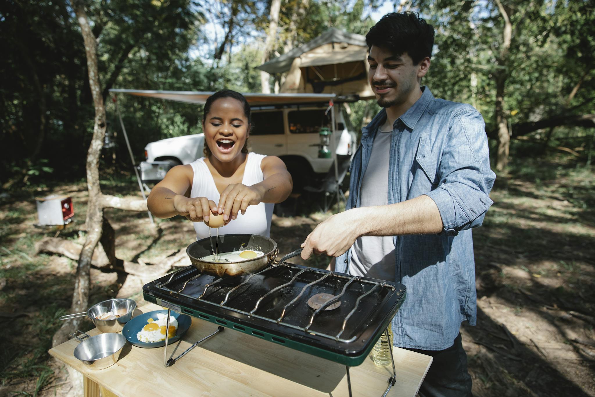 High angle of positive multiracial couple frying eggs on skillet using metal gas stove on wooden table near car and tent in forest