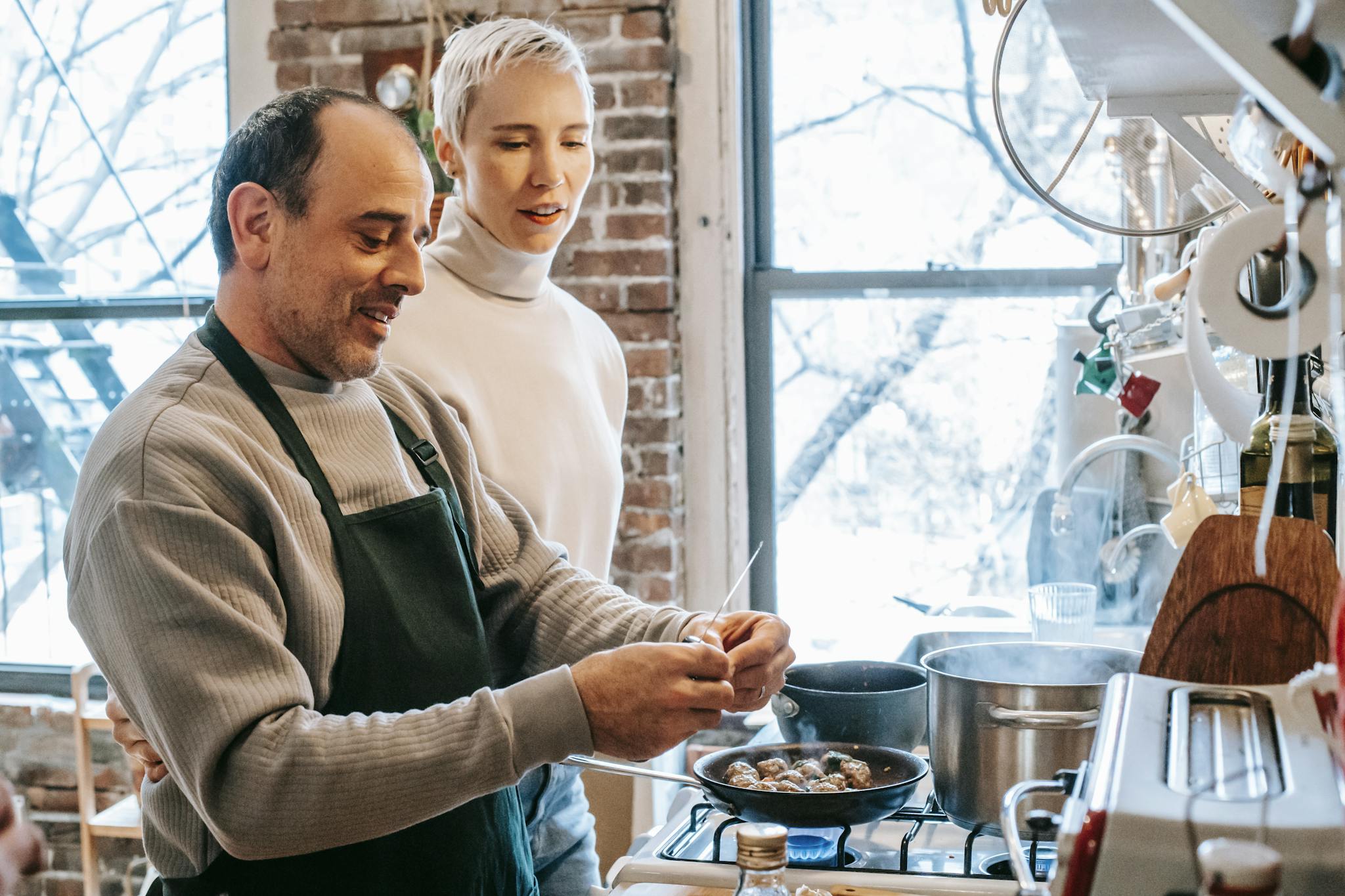 Smiling man in apron cooking against attentive girlfriend and gas stove with meatballs in pan at home